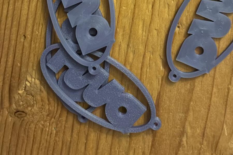 Tre portachiavi DMH stampati in 3D, color lavanda, appoggiati su un tavolo di legno